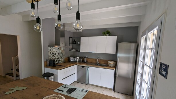 Eine moderne Küche mit weißen Schränken, einem Edelstahlkühlschrank und einer Holzarbeitsplatte. Hängelampen und eine Pflanze sind an der Wand angebracht.