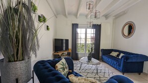 Ein Wohnzimmer mit einem blauen Sofa, einer großen Kübelpflanze, einem Fernseher und einem Esstisch mit Stühlen.