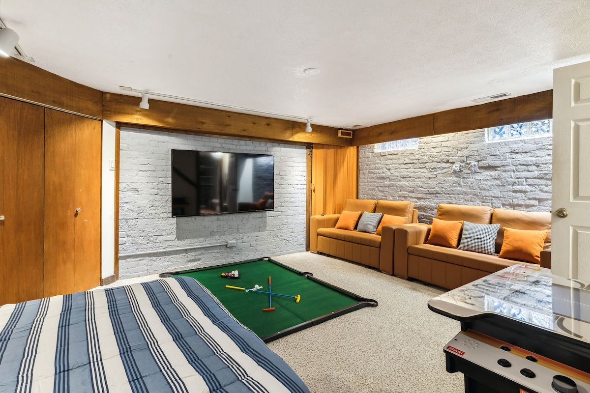 Una habitación con mesa de billar, un conjunto de sofás, un televisor montado en una pared de piedra y paneles de madera.