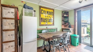 Una cocina con un refrigerador blanco, una mesa de madera y sillas, un letrero amarillo en la pared y un bote de basura verde.