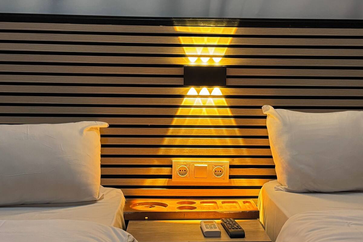 A hotel room with a wooden headboard, two white pillows, a bedside table with a remote control, and a wall-mounted light fixture.