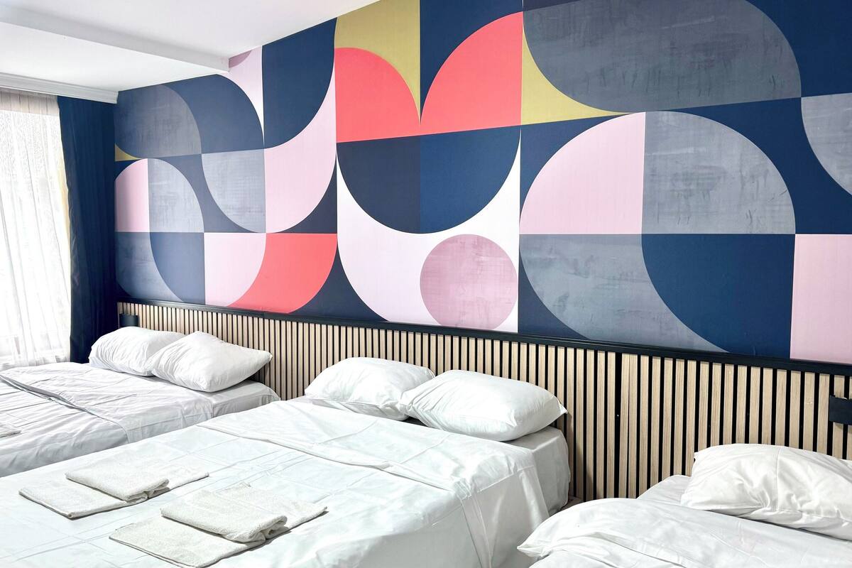 A hotel room with two beds, a large abstract wall mural, and a window with sheer curtains.