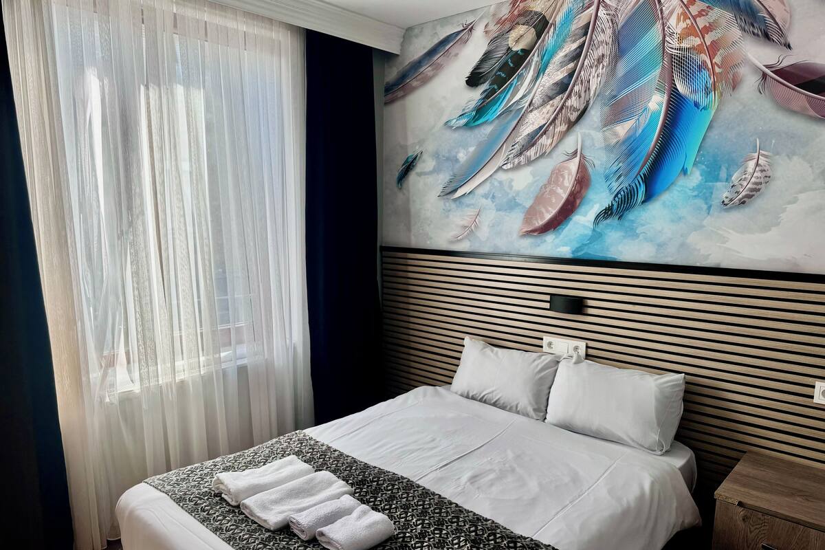 A hotel room with a bed, a wall mural of feathers, a window with sheer curtains, and a bedside table.