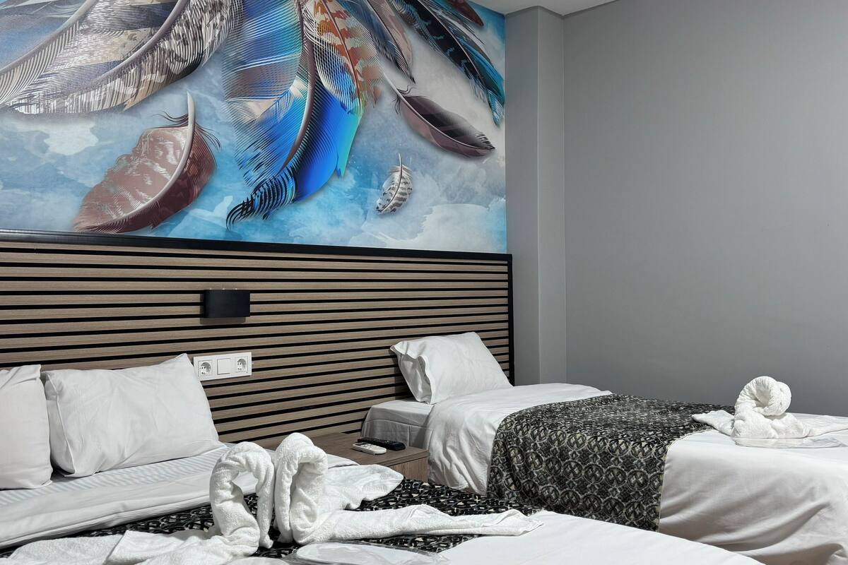 A modern hotel room with a large feather mural on the wall, two beds with white linens, and a bedside table with a remote control.