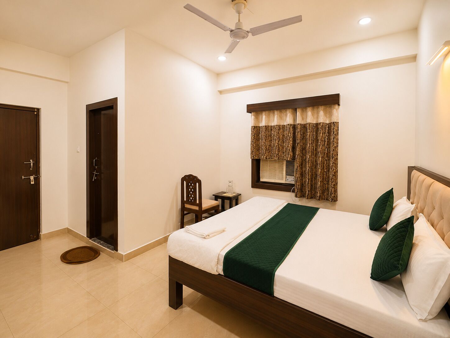 Une chambre d’hôtel avec un lit, une chaise, un bureau, une fenêtre avec des rideaux, un ventilateur de plafond et une porte.