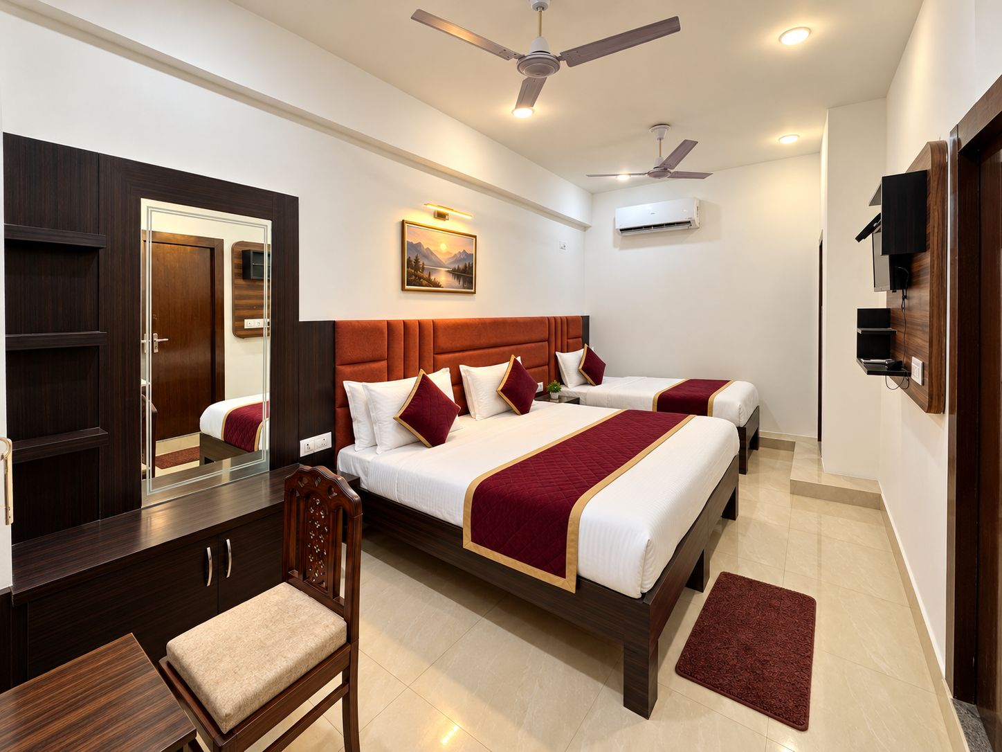 A hotel room with two beds, a desk, a chair, a mirror, ceiling fans, and an air conditioning unit.