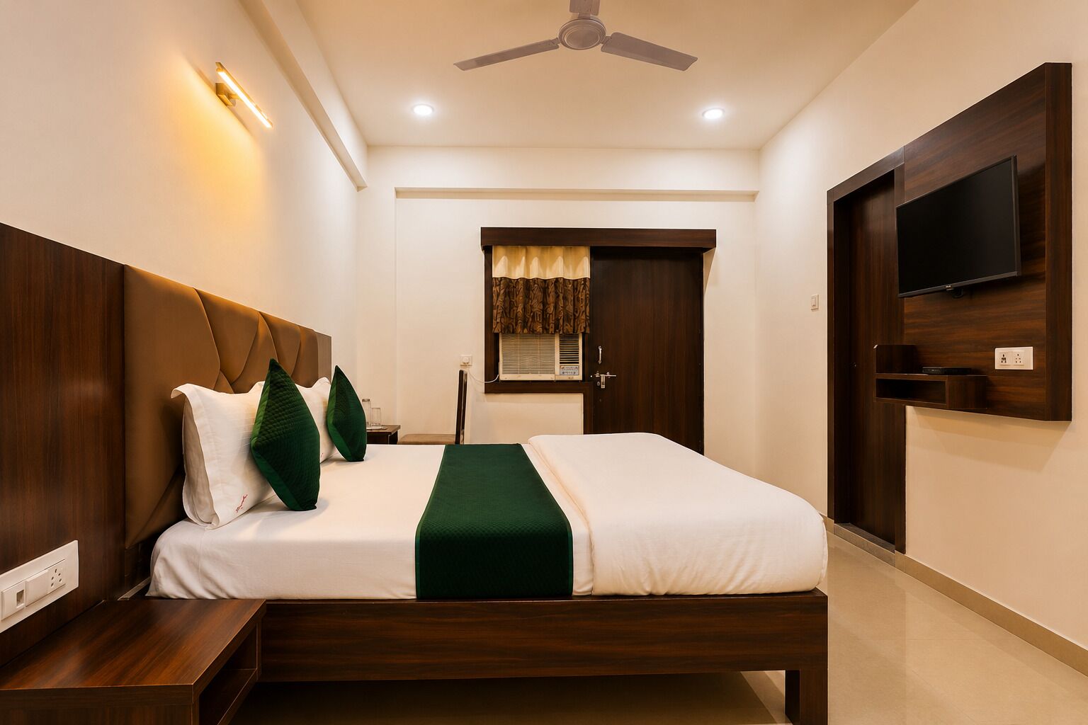Habitación de hotel con cama, mesitas de noche, ventilador de techo, televisor montado en la pared y ventana con cortinas.