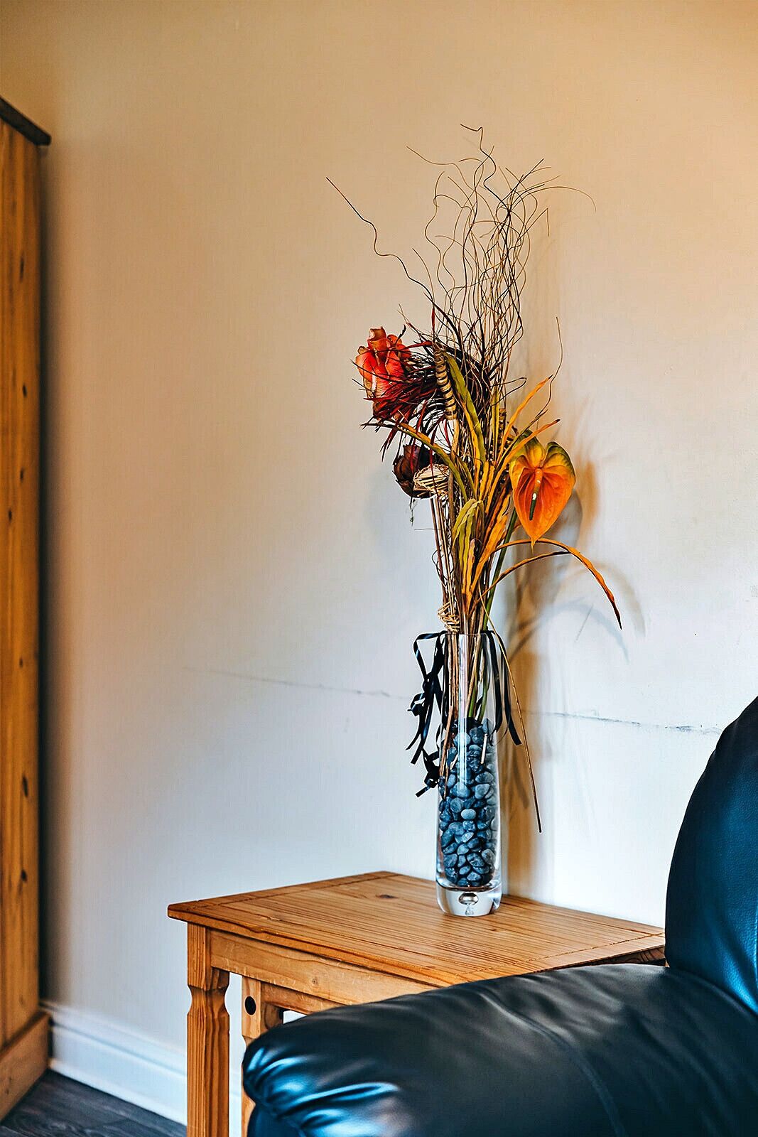 A vase with dried flowers and a single blooming flower on a wooden table against a plain wall.