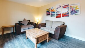 Een woonkamer met een bruine leren bank, een grijze bank, een houten salontafel en een kleine houten bijzettafel. Op de muur hangen drie abstracte schilderijen.