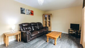 A living room with a leather sofa, wooden coffee table, a TV stand, and a bookshelf. - Camelia Country Cottage (Dorrington)