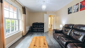 A living room with a leather sofa, a wooden coffee table, and a view of a garden through large windows. - Camelia Country Cottage (Dorrington)