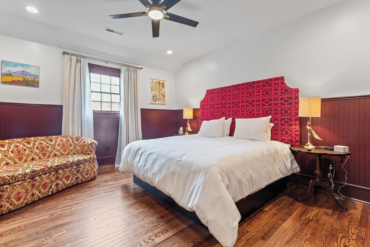 Quarto de hotel com uma cama grande, um sofá, piso de madeira, ventoinha no teto e uma janela com cortinas.