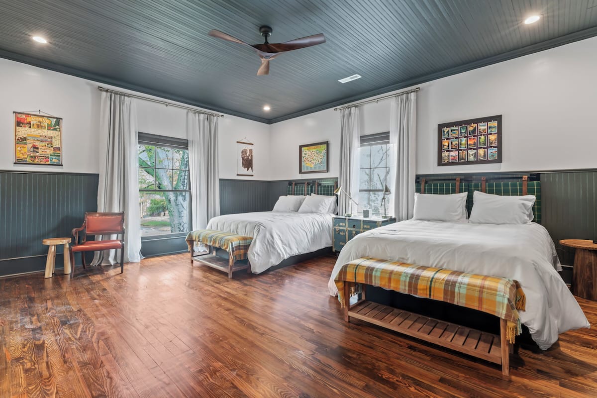 Quarto de hotel com duas camas, ventoinha no teto, soalho de madeira e janelas amplas com cortinas.