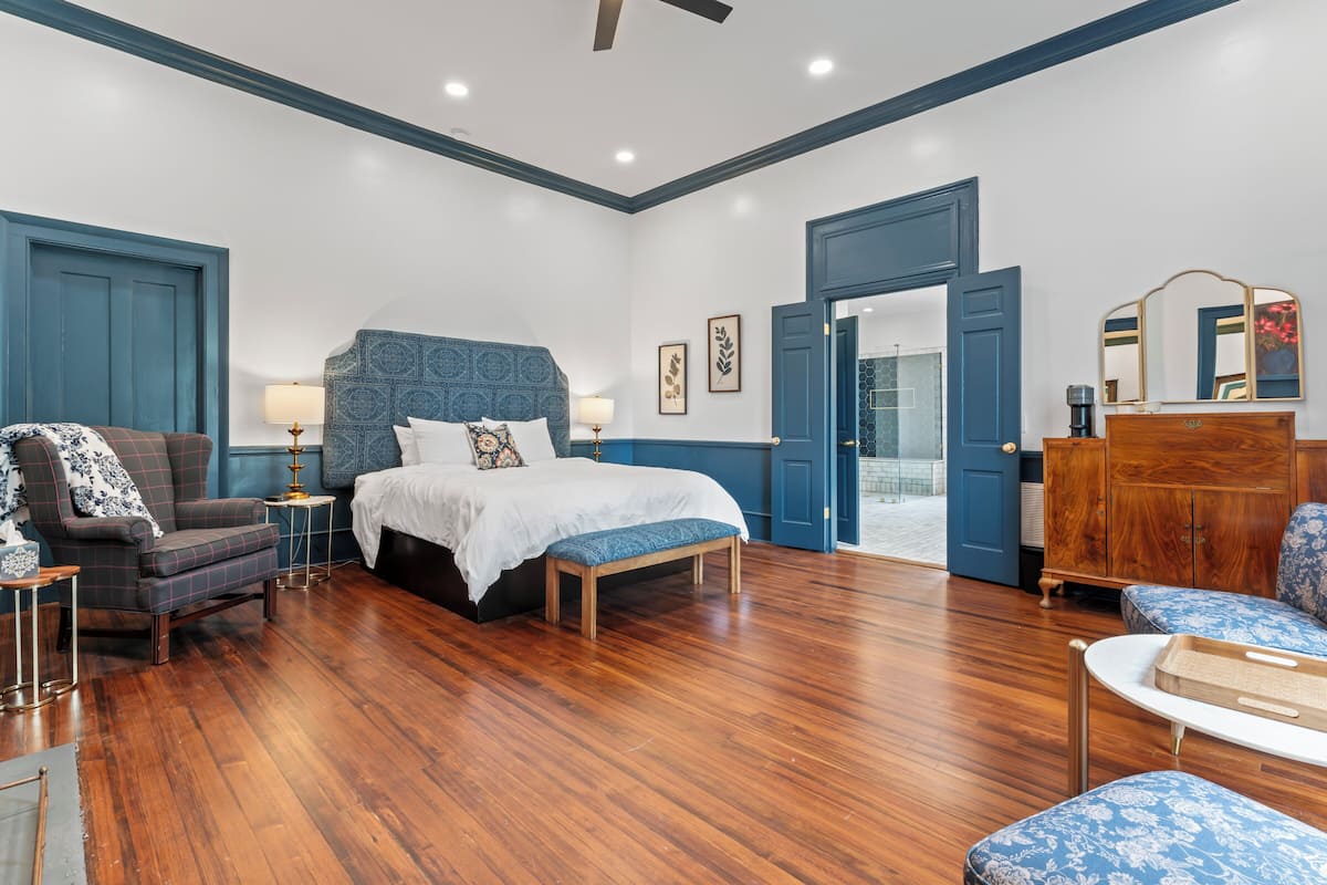 Quarto com cama, uma cadeira, um banco, cômoda e porta que dá para outro ambiente.
