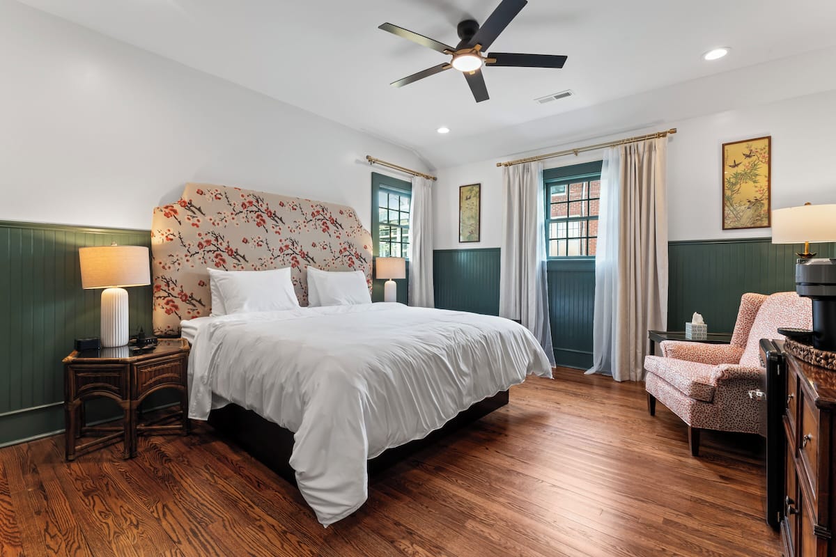 Quarto de hotel com uma cama grande, ventoinha no teto, mesa de cabeceira com abajur, cadeira e janela com cortinas.