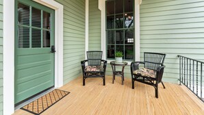 A porch with two wicker chairs and a small table, a green door, and a window with a view of the backyard.