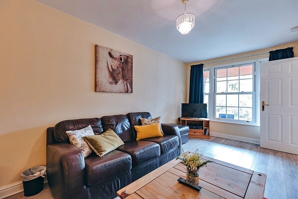 A living room with a leather sofa, a coffee table with a vase, a television on a stand, and a large window with curtains. - Peach Tree County Cottage (Dorrington)