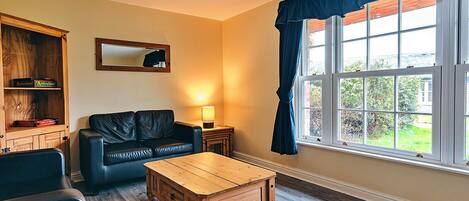 Una sala de estar con un sofá de cuero negro, una mesa de centro de madera, un aparador con libros, una lámpara y un ventanal con cortinas azules.