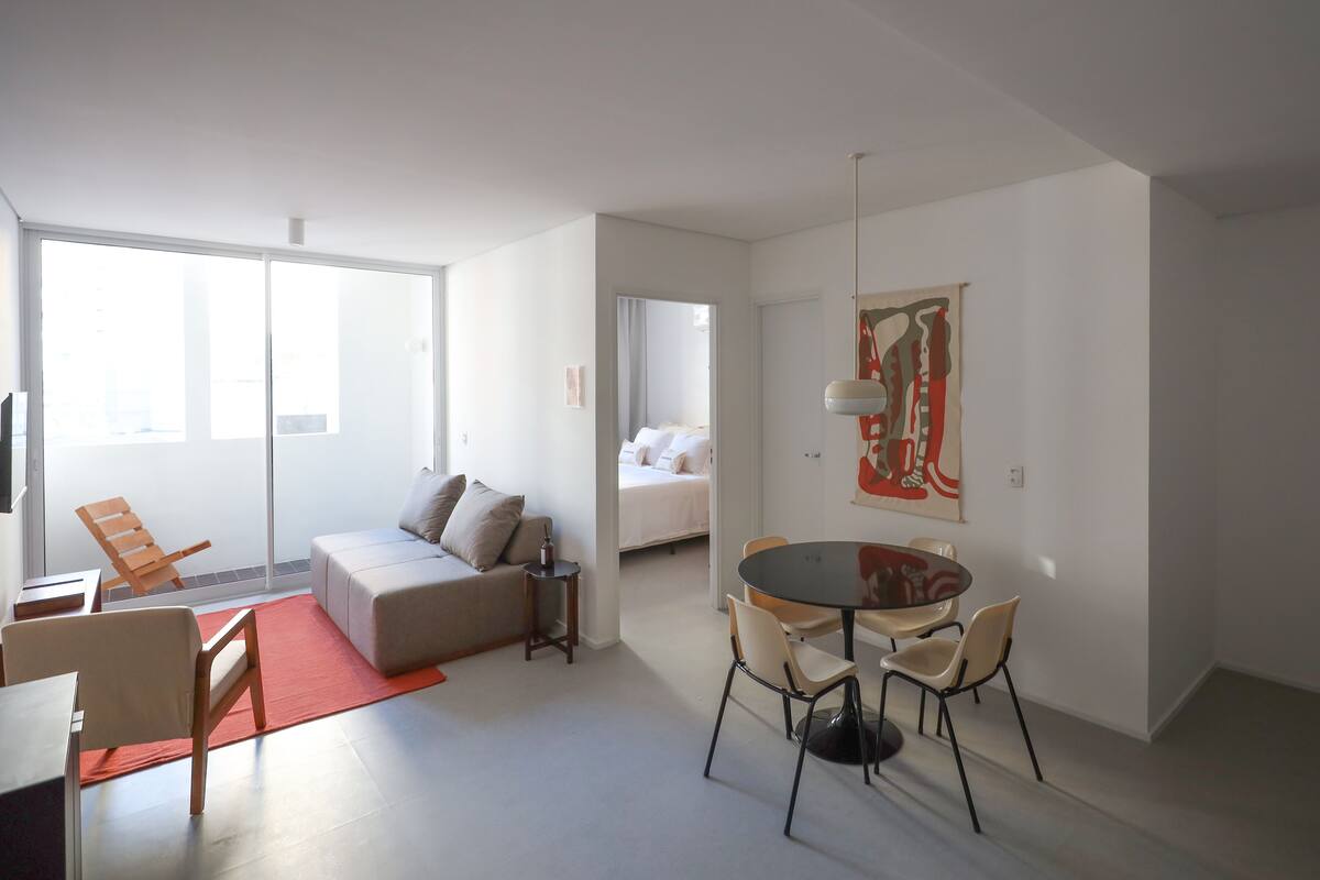 Ein modernes Wohnzimmer mit einer Couch, einem Esstisch mit Stühlen, einem kleinen Beistelltisch und einem großen Fenster.