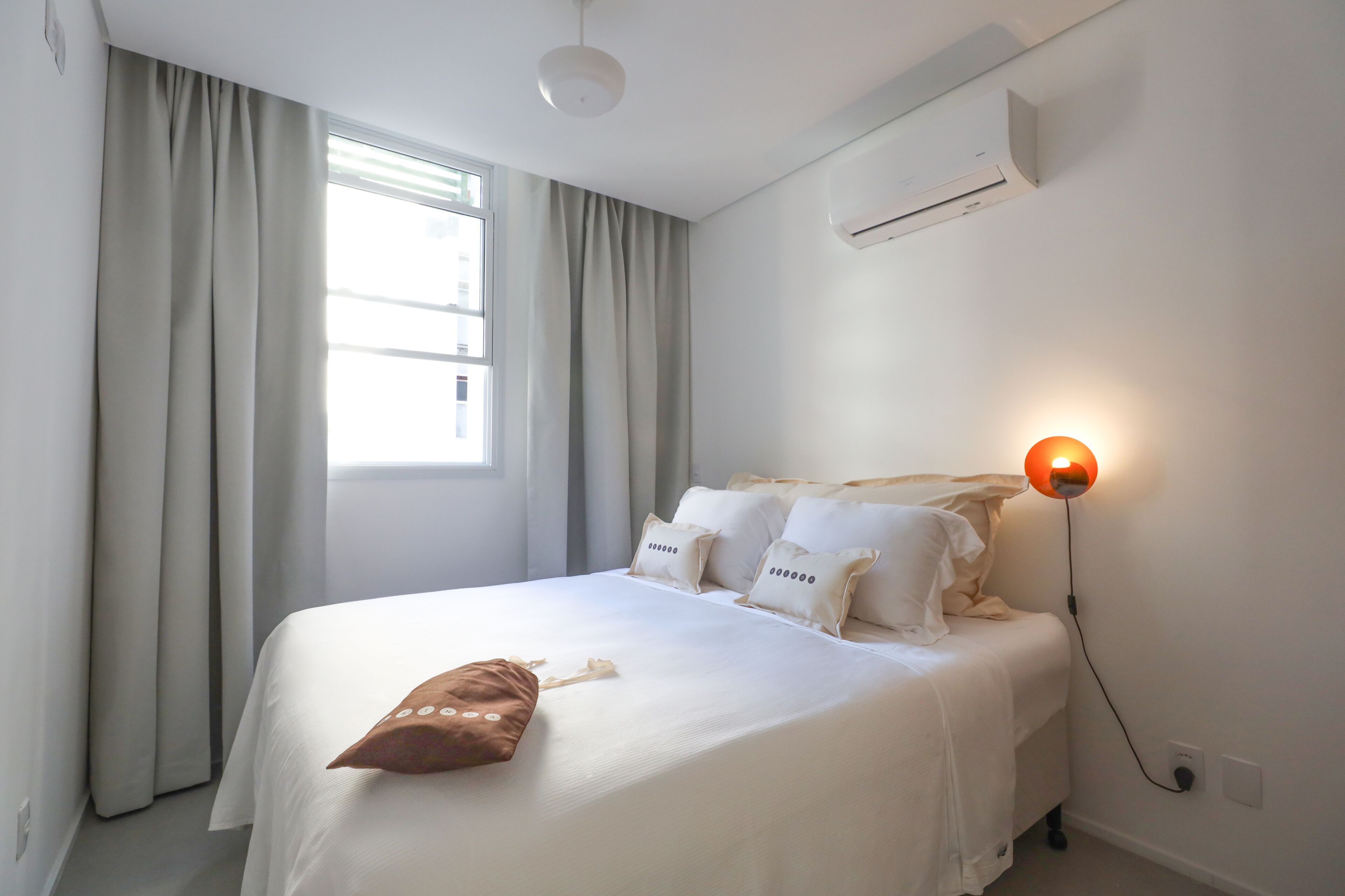 Ein ordentlich gemachtes Bett mit weißer Bettwäsche und mehreren Kissen, einer Nachttischlampe und einem Fenster mit Vorhängen.
