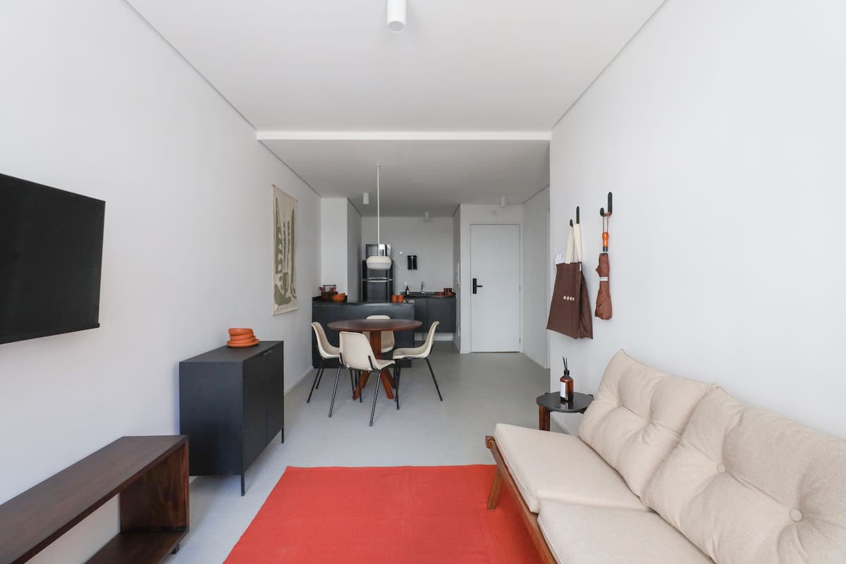Ein modernes Wohnzimmer mit einer Couch, einem Esstisch mit Stühlen, einem an der Wand montierten Fernseher und einer Küche im Hintergrund.