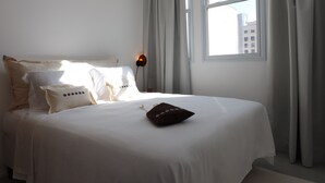 A neatly made bed with white bedding and a single brown pillow, a bedside lamp, and a window with curtains.