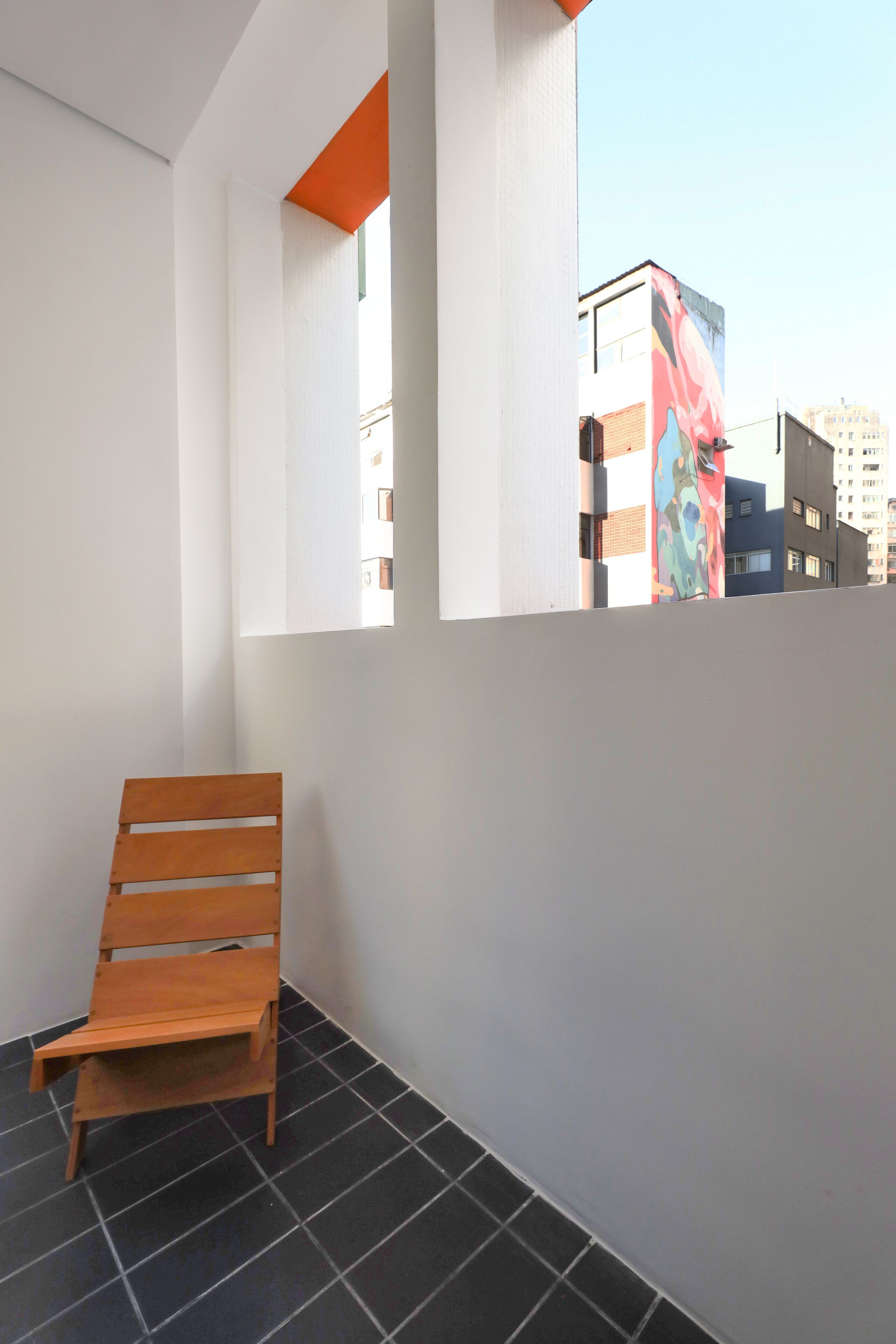 Een balkon met een houten stoel, witte muren en uitzicht op gebouwen.