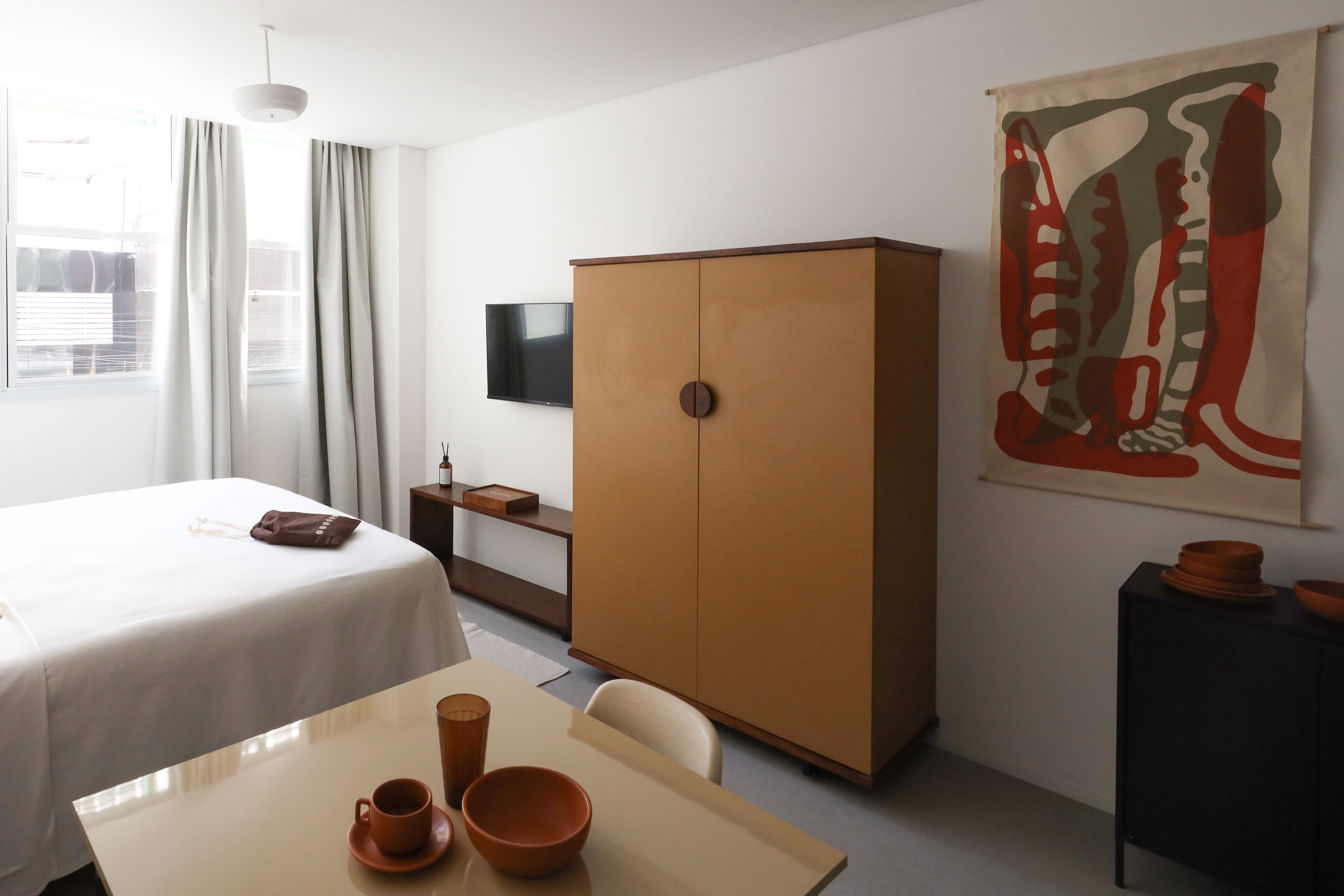 Ein Hotelzimmer mit einem Bett, einem Tisch mit Tasse und Schüssel, einem Kleiderschrank, einem Fernseher und einer Wand mit einem großen abstrakten Gemälde.