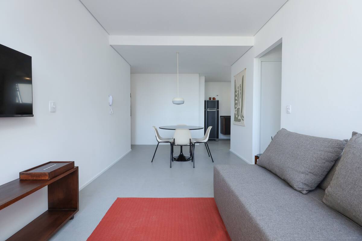 Ein modernes Wohnzimmer mit grauem Sofa, einem roten Teppich, einem Esstisch mit Stühlen und einem an der Wand befestigten Fernseher.