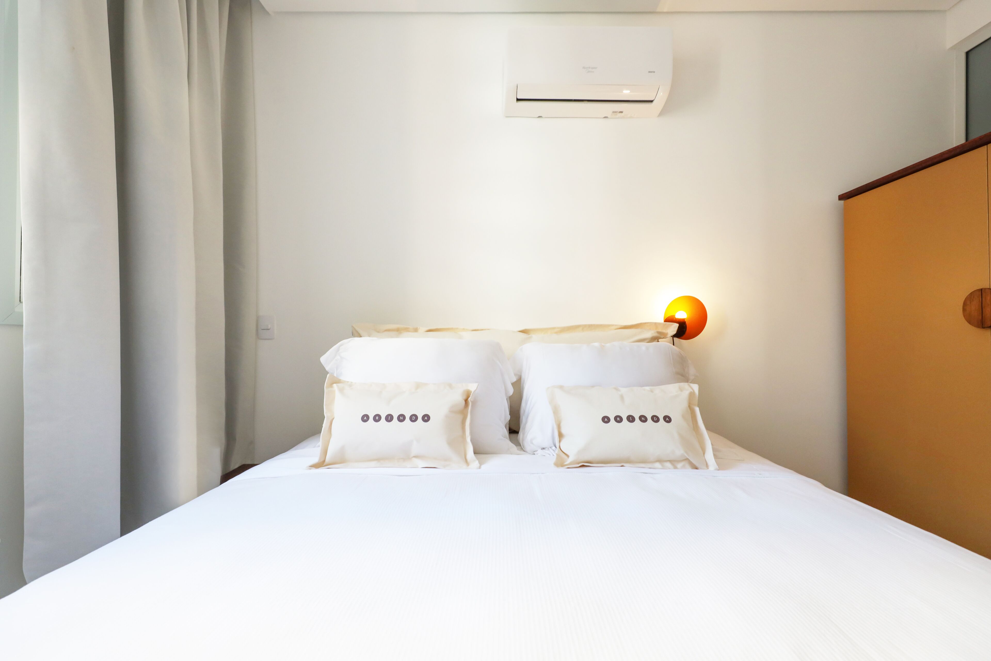 Une chambre d’hôtel avec un lit, du linge de lit blanc, un climatiseur fixé au mur et une lampe de chevet.