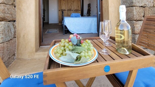 Un balcon avec une table où sont disposés une assiette de raisins et une bouteille de vin, un lit recouvert d’une couverture bleue et un mur en pierre.