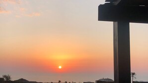 Un coucher de soleil sur un paysage désertique, avec une végétation clairsemée et une construction visible au premier plan.