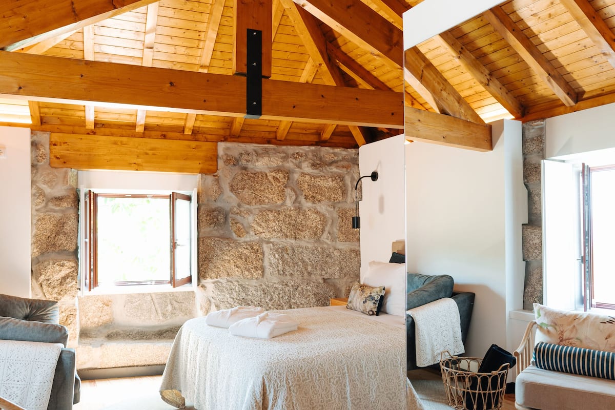 Ein Schlafzimmer mit einer Steinwand, Holzbalkendecke, einem Bett, einem Sessel und einem Fenster.