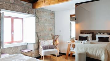 A bedroom with a wooden ceiling, stone walls, a large bed, a bedside table, a chair, and a window with curtains.