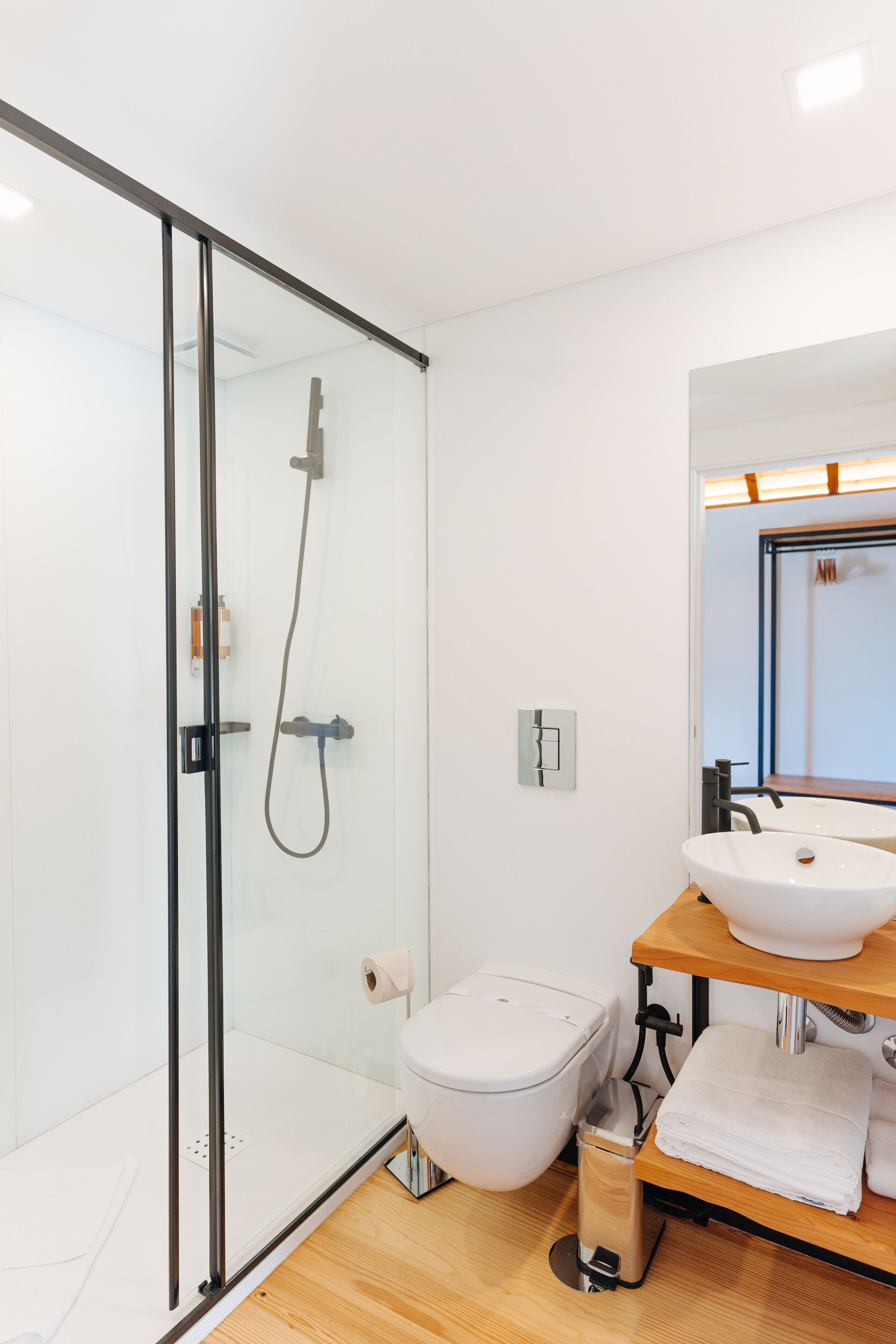 Et moderne badeværelse med en glasbrusekabine, et hvidt toilet, en trævæg med vask og et spejl.