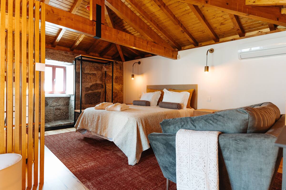 Ein Schlafzimmer mit einem Bett, einer grauen Couch, einer Holzbalkendecke und einer Steinwand.