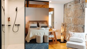 A modern bedroom with a stone wall, wooden ceiling beams, a bed with white bedding, a small seating area, and a shower. - D'Autor Village (Baiao)