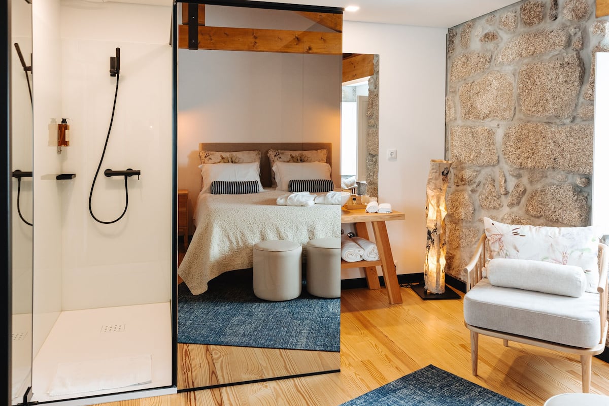 Ein modernes Schlafzimmer mit Steinwand, Holzbalkendecke, einem Bett mit weißer Bettwäsche, einer kleinen Sitzecke und einer Dusche.