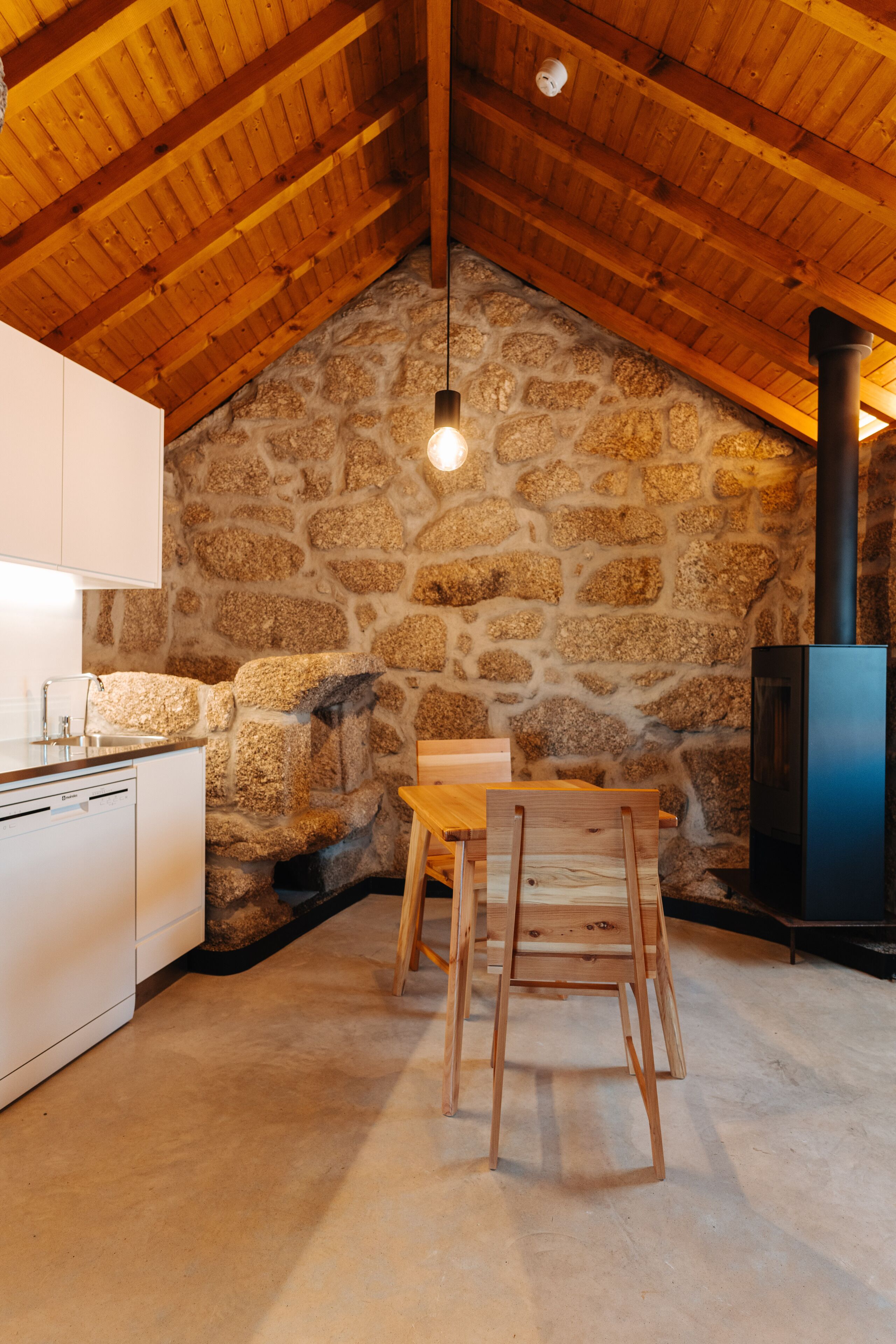 A kitchen with a stone wall, wooden ceiling beams, a wooden table, and a black stove.