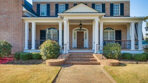 A two-story brick house with a front porch, a central door, and a small front yard with a brick pathway and two bushes. - Riverfront North Augusta Home w/ Private Pool! (North Augusta)