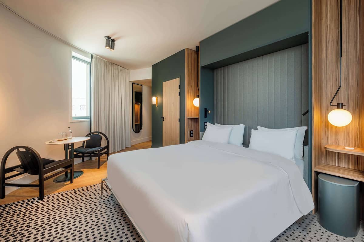 Une chambre d’hôtel avec un grand lit, un bureau avec deux chaises, une petite table avec une chaise et une fenêtre avec des rideaux.
