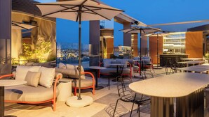 Un espacio al aire libre con sillas acolchadas, mesas y sombrillas, con vistas a un paisaje urbano.
