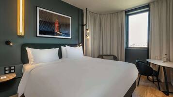 A hotel room with a large bed, a desk, and a chair. There is a window with curtains, a wall-mounted light, and a framed picture on the wall.