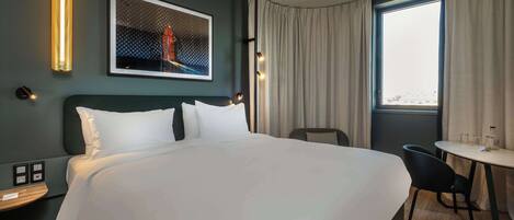 Quarto de hotel com uma cama grande, uma escrivaninha e uma cadeira. Há uma janela com cortinas, uma luminária na parede e um quadro emoldurado.