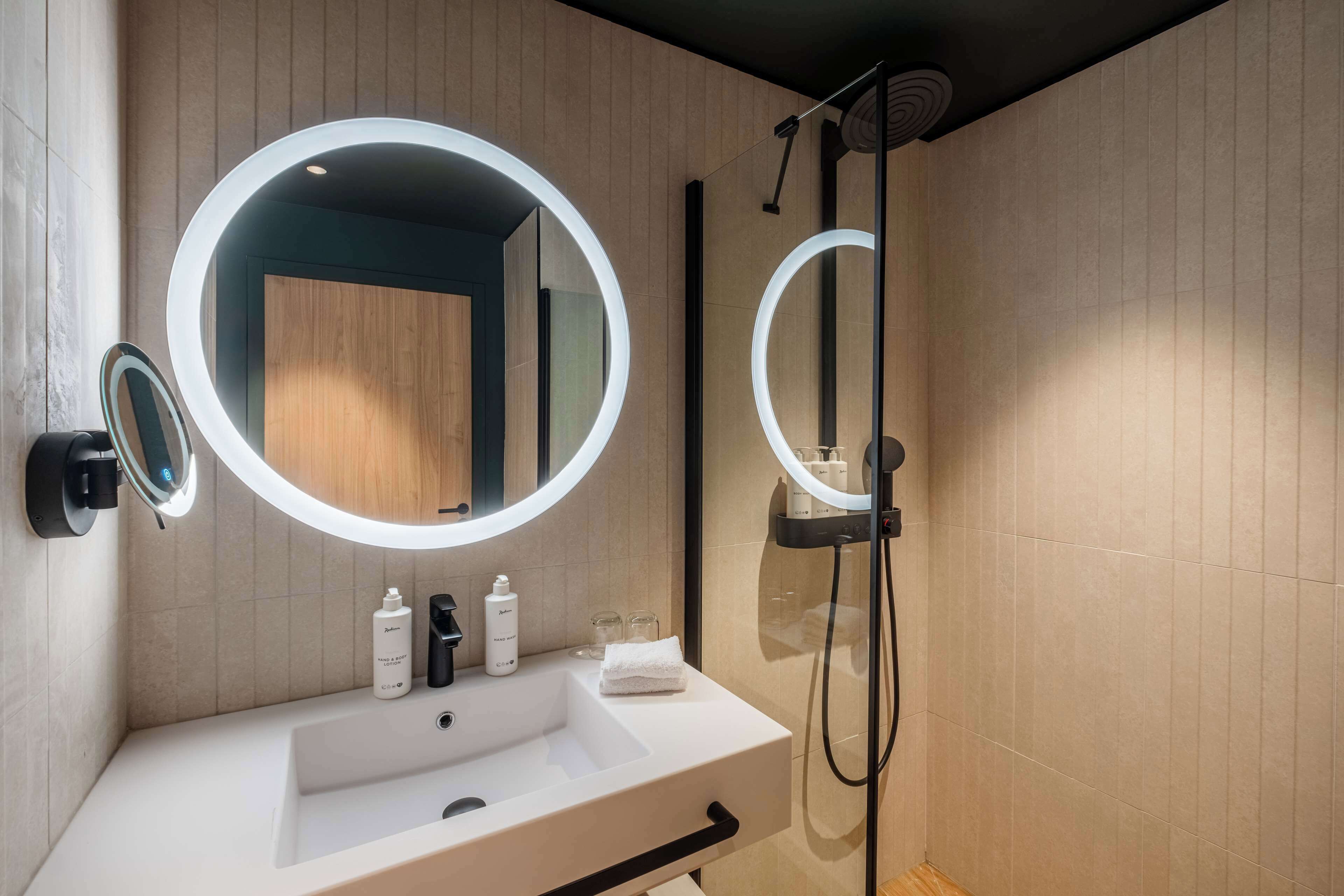 A modern bathroom with a white sink, two round mirrors with lights, a handheld showerhead, and a towel on the counter.