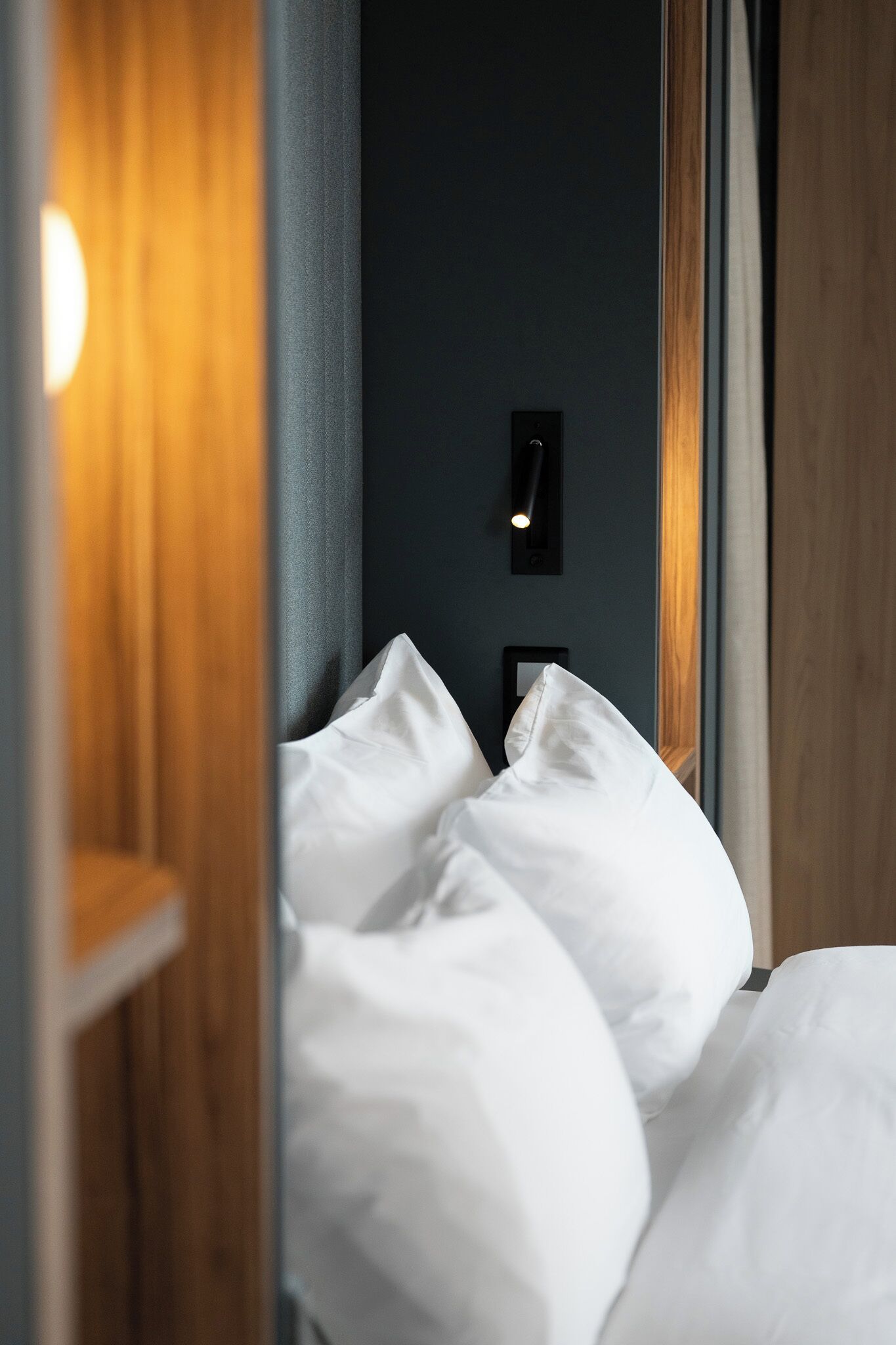 Una cama bien hecha con sábanas blancas, una pared oscura y una pequeña lámpara negra arriba.