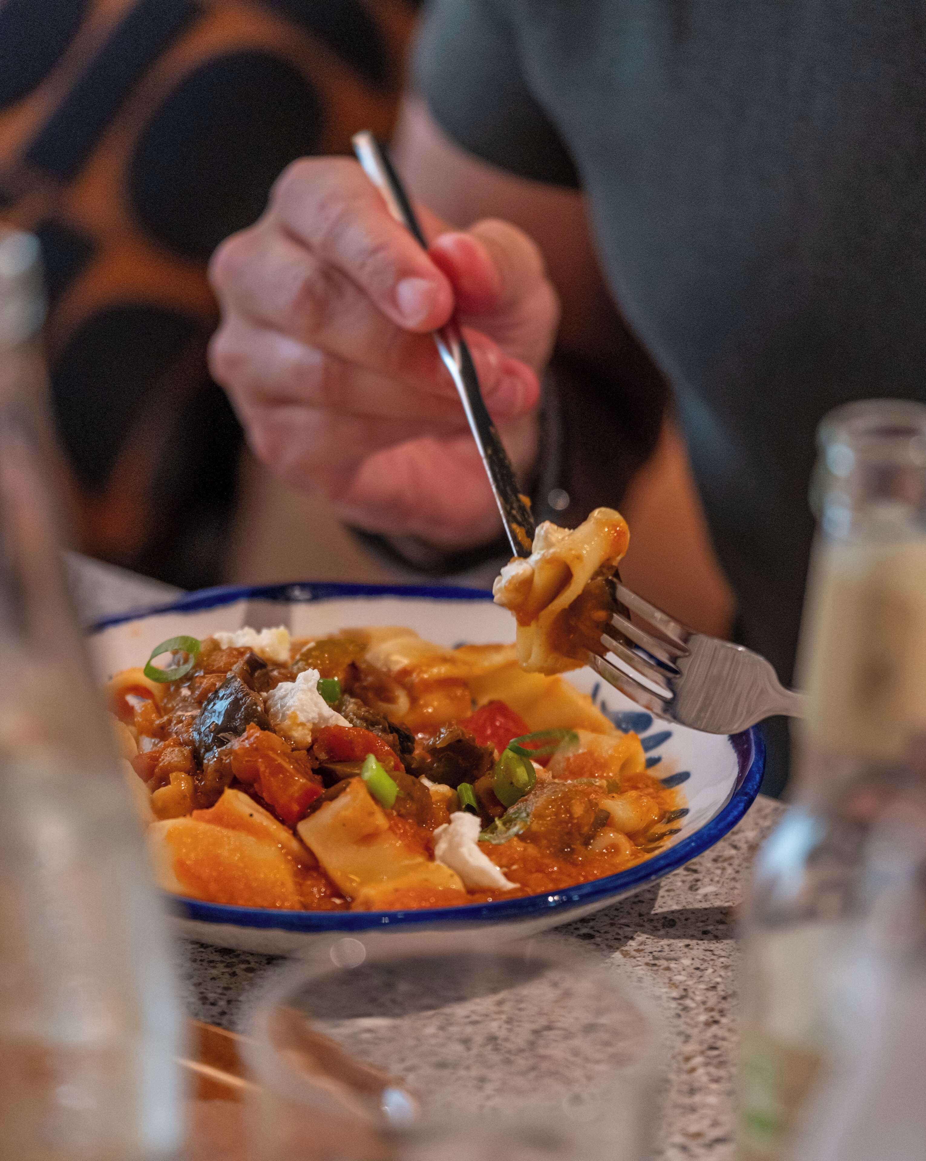Una persona usando un tenedor para comer un plato de pasta, verduras y salsa cremosa.