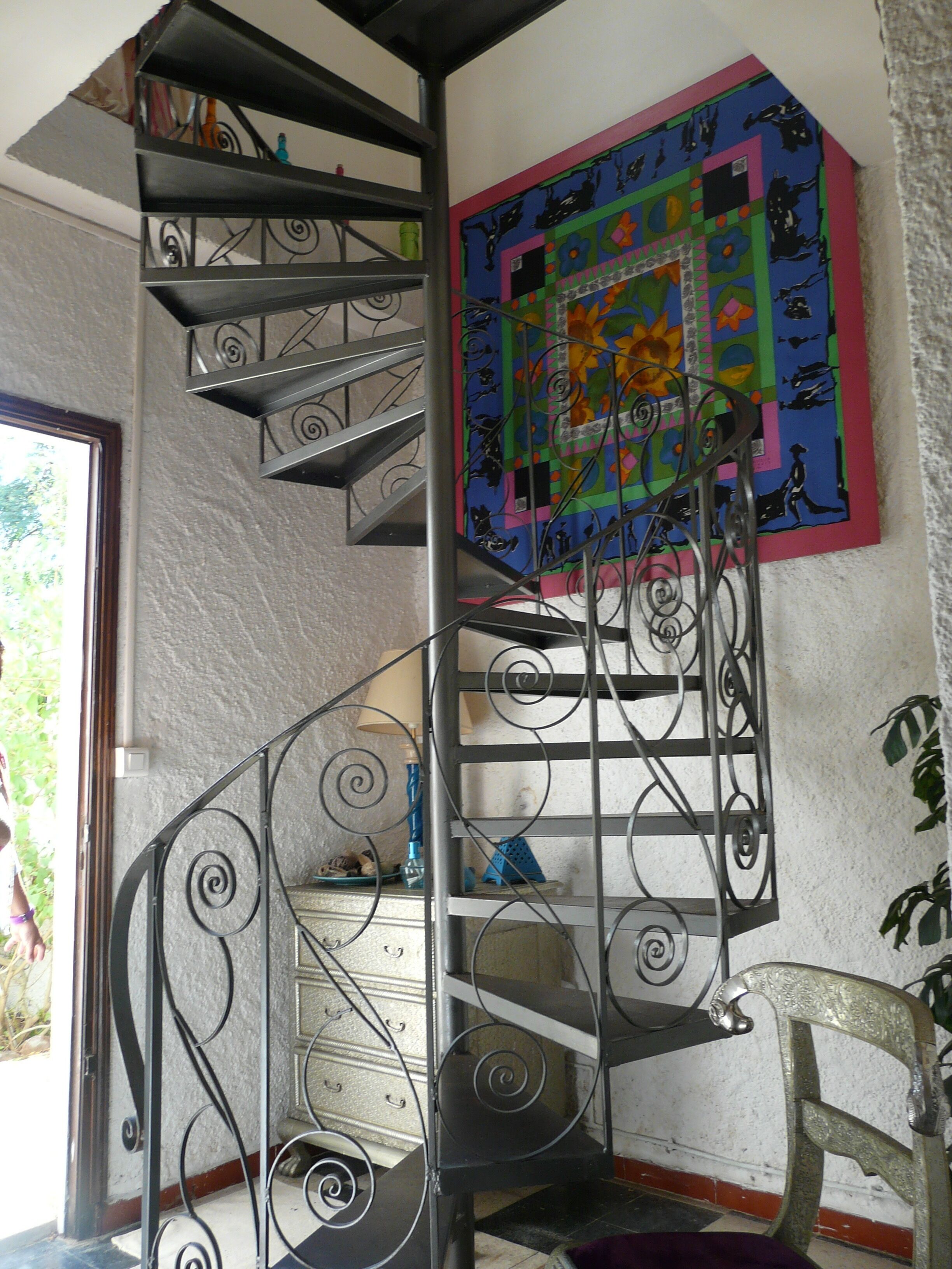 Un escalier en colimaçon avec une rampe en métal, une fresque murale colorée et une chaise dans une pièce dont le mur est texturé et qui possède une fenêtre.