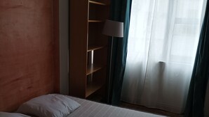 A bedroom with a bed, pillows, a wooden shelf, a floor lamp, and a window with curtains. - Parisian apartment (Paris)
