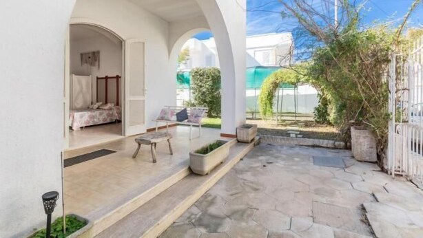 A courtyard with a stone floor, a covered seating area, and a white building with an archway leading to a room with a bed and a window. - Villa Biancarosa - Rosa by BarbarHouse (Porto Cesareo)